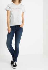 Camiseta de algodón gris con el logotipo de "Tommy Jeans", combinada con jeans skinny azul oscuro y zapatillas negras con acentos blancos. Un atuendo simple y casual.