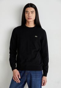 Sort strikket sweater med lange ærmer, rund hals og ribbede manchetter. Har et lille grønt Lacoste-logo på brystet.