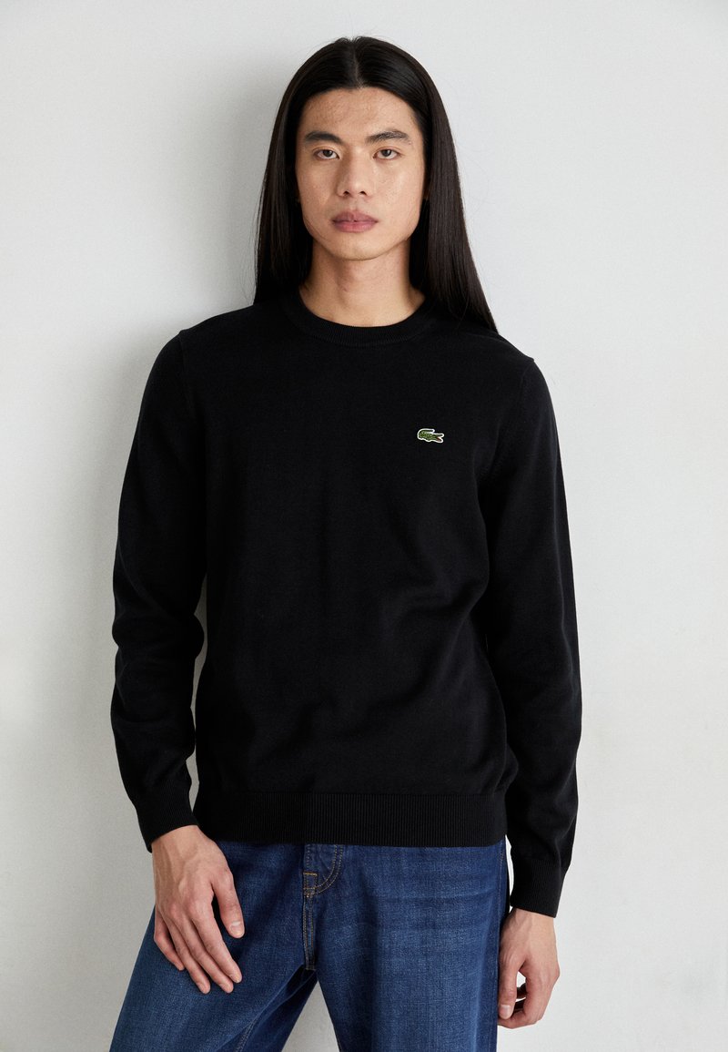 Sort strikket sweater med lange ærmer, rund hals og ribbede manchetter. Har et lille grønt Lacoste-logo på brystet.