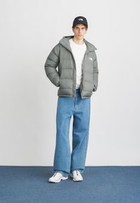 Szürke steppelt kabát kapucnival, fehér póló, világoskék bővülő nadrág és fehér sportcipő. Fekete sapka elől logóval.