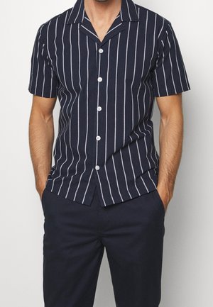 Mann trägt ein marineblaues kurzärmeliges Hemd mit weißen vertikalen Streifen und dunkelmarineblaue Hose, Hände in den Taschen, vor einem schlichten Hintergrund.