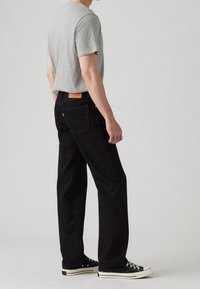 Personne portant un jean noir à coupe droite, un t-shirt gris à manches courtes et des baskets noires basses, debout devant un fond clair uni.