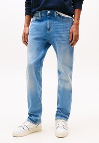 Mees, kes kannab helesiniseid kulunud teksapükse ja valgeid tossusid, seisab käed lõdvalt külgedel tühja tausta ees.