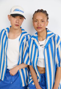 Blauw en beige gestreepte overhemden met korte mouwen, gemaakt van een gestructureerde stof, gecombineerd met een witte tanktop en blauwe onderkleding. Een baseballcap is zichtbaar.