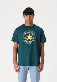 Camiseta de algodón color teal con un gráfico circular amarillo de "Converse All Star" con una estrella en el centro. Cuello redondo clásico y mangas cortas.