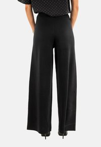 Pantaloni neri a gamba larga realizzati in un tessuto liscio, dotati di una vita elasticizzata. La vista posteriore mostra un taglio a lunghezza intera e un design minimalista.