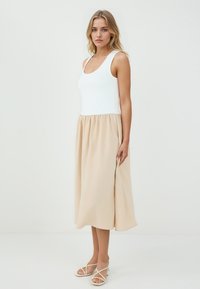 Un top blanc sans manches associé à une jupe beige, longueur genou. La jupe a une taille froncée et une texture lisse. Des sandales blanches à lanières complètent le look.