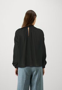 Blusa nera con collo alto, schiena arricciata con dettaglio a chiave e lunghe maniche ampie. Abbinata a pantaloni a gamba larga azzurri.