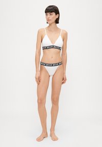 Bijela trokutasta braleta i odgovarajuće bikini gaćice s crnim elastičnim trakama na kojima je ispisan tekst "justcavalli". Glatka tekstura, minimalistički dizajn.