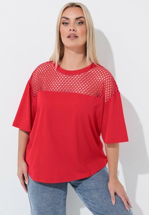 Frau mit glatten blonden Haaren, die ein rotes Oberteil mit durchscheinendem oberen Bereich und blaue Jeans trägt, steht vor einem weißen Hintergrund.