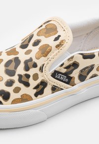 Sneaker slip-on Vans in tela beige con stampa leopardata oro e nera, suola in gomma bianca, accenti elastici laterali e etichetta con logo.