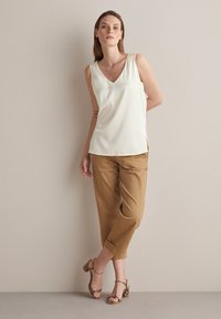 Biała bluzka bez rękawów w szpic, zestawiona z jasnobrązowymi spodniami 3/4 i wzorzystymi obcasami, prezentowana na neutralnym tle.