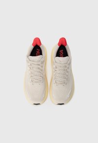 Baskets de sport beiges avec un dessus texturé, accents rouges sur l'onglet du talon et semelle jaune. Comprend un design à lacets et des détails réfléchissants.