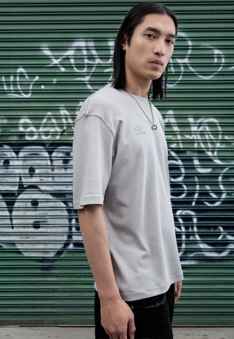 T-shirt de manga curta cinza claro com um ajuste ligeiramente solto, apresentando uma marca discreta. Usada com calções escuros contra um fundo de graffiti.