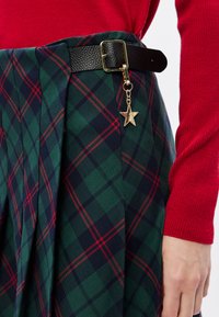 Μαύρη ψεύτικη δερμάτινη ζώνη με χρυσό κούμπωμα και γοργόνι, συνδυασμένη με πλισέ φούστα καρό σε πράσινη και ναυτική απόχρωση με κόκκινες λεπτομέρειες.