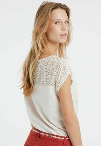 Crème kortarmige top met een kant achterpaneel, een gladde voorkant en een ontspannen pasvorm. Gedragen met rode broeken en een gestreepte riem.