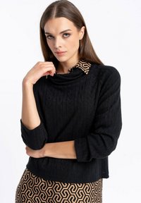 Czarny sweter dzianinowy z teksturowanym wzorem, z dekoltem typu cowl i rękawami do trzech czwartej. W połączeniu z brązową spódnicą w geometryczny wzór.