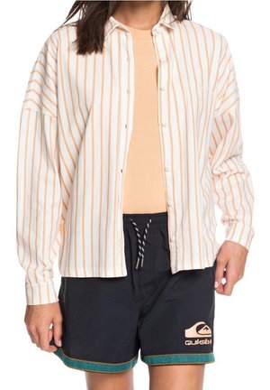 Osoba nosząca beżową koszulę w paski z guzikami nałożoną na brzoskwiniowy T-shirt oraz czarne szorty Quiksilver z turkusowym wzorzystym obszyciem.