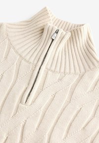 Suéter de punto de color beige claro con un alto cuello acanalado, que presenta un cierre de media cremallera y un patrón de cable texturizado en toda su extensión.