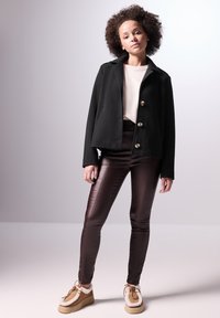 Giacca nera corta con chiusura a bottoni, maglione crema, pantaloni marroni aderenti e scarpe marroni chiare con dettagli in pelliccia. Tessuti morbidi e vestibilità su misura.