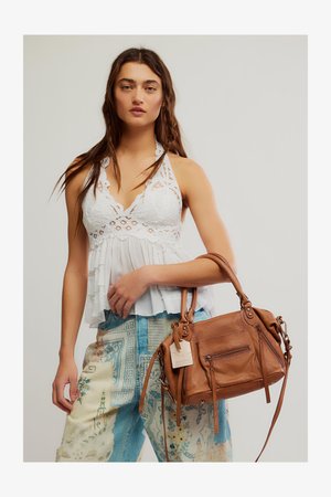 Donna con lunghi capelli castani indossa un top all'uncinetto bianco con collo a halter e pantaloni fantasia, tenendo una borsa in pelle marrone con etichette.