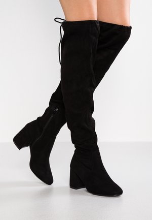 Jambes portant des bottes noires en daim au-dessus du genou avec des talons blocs et des fermetures éclair sur le côté intérieur, posées sur une surface blanche et lisse.