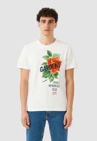 T-shirt bianco in cotone con una grande grafica di una rosa arancione, foglie verdi e la parola "GIARDINI" in grassetto nero, con testo stampato sottostante.