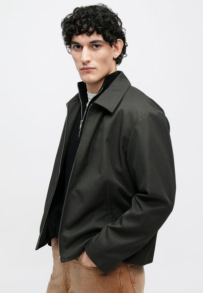 Tmavosivá bunda s golierom, zapínaním na zips a bočnými vreckami. Vyrobená z hladkého materiálu, s voľným strihom a minimalistickým dizajnom.