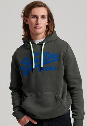 Groene hoodie met blauwe "SuperDry" tekst en logo. Voorzien van een voorzak en mintgroene trekkoorden. Zachte stof met een relaxte pasvorm.