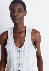 Lys blå denimvest med scoop-hals, frontlukking med knapper og uten ermer. Har en tettsittende fasong og subtile sømdetaljer.
