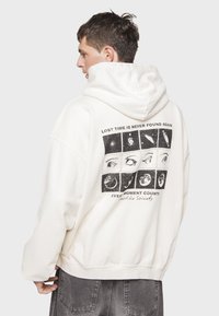 Sudadera de algodón blanca con un estampado gráfico en la parte posterior, que presenta paneles negros con imágenes de ojos. Puños acanalados y capucha con cordón.