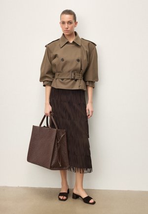 Femme portant une veste olive ceinturée et une jupe plissée marron foncé, tenant un grand sac tissé marron et portant des sandales à talon bloc ouvertes.