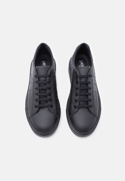 Paire de baskets basses noires avec un motif en relief sur les côtés et des lacets noirs, photographiées de dessus sur un fond blanc.