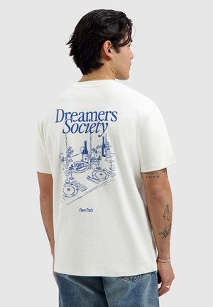 Man with dark hair wearing white T-shirt showing blue "Dreamers Society" text and dining table illustration on back.