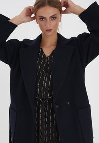 Navy-Wollmischungsjacke mit strukturiertem Design, Reverskragen, doppeltknopf und Seitentaschen, getragen über einem gemusterten schwarzen Kleid.
