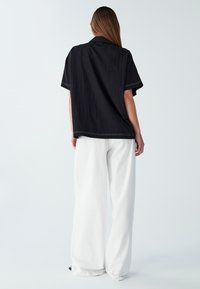 Czarna koszulka z krótkim rękawem o fakturowanym wzorze, z białymi detalami wykończenia; w połączeniu z luźnymi, białymi spodniami typu wide-leg. Model widziany z tyłu.