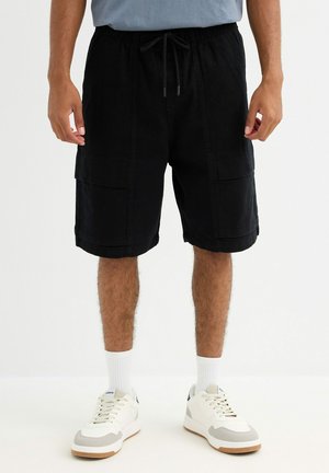 Hombre con pantalones cortos cargo negros, calcetines blancos hasta la mitad de la pierna y zapatillas blancas sobre un fondo claro y liso.