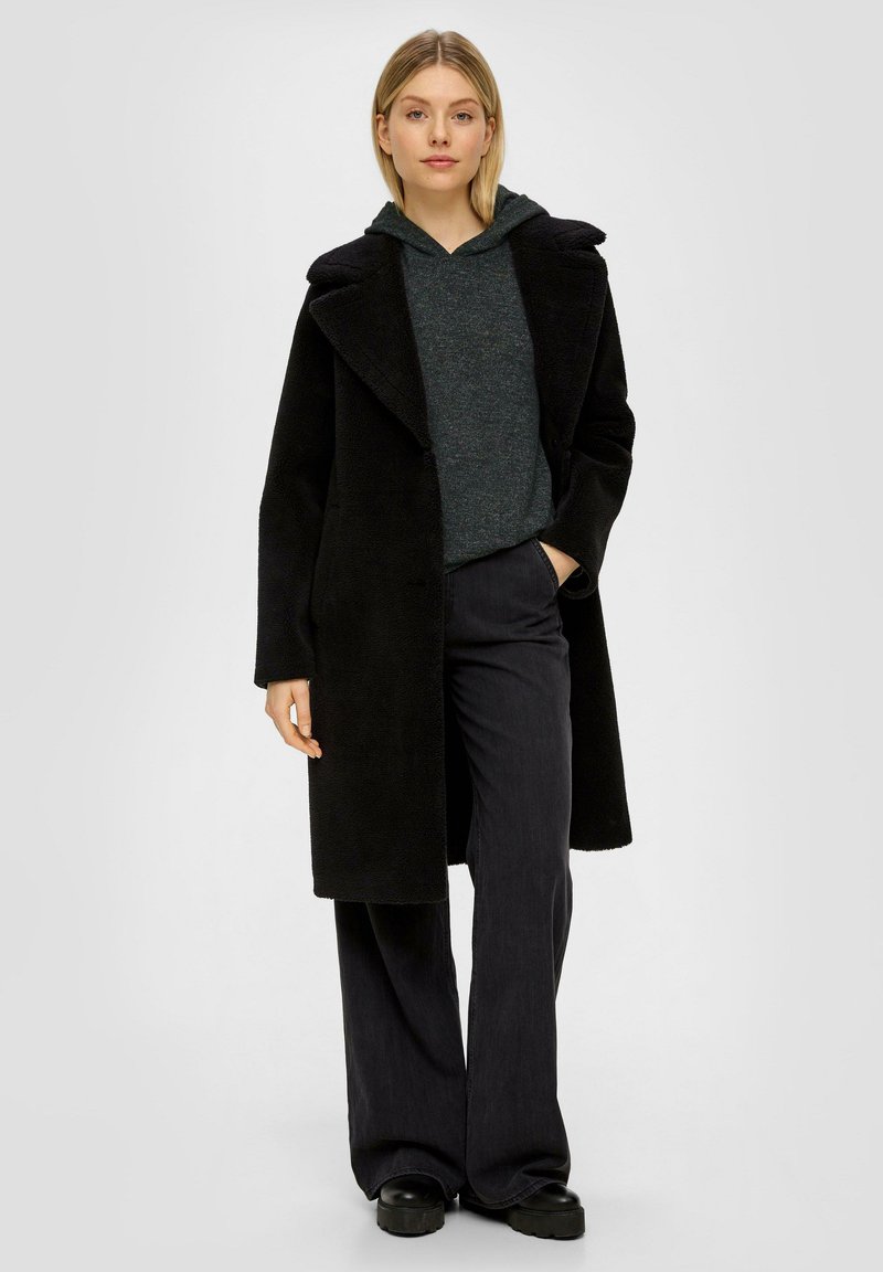 Schwarzer Wollmantel mit breiten Revers, getragen über einem dunkelgrauen Kapuzenpullover. Schwarze weit geschnittene Hose und schwarze Plateauschuhe runden das Outfit ab.