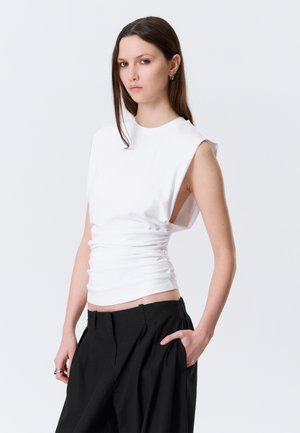 Jeune femme aux longs cheveux bruns portant un haut blanc sans manches froncé et un pantalon noir à jambes larges, debout avec une main dans la poche.