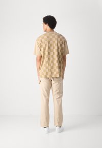 Camiseta corta de manga en color beige con un patrón de tablero de ajedrez y detalles de logo, combinada con pantalones cargo beige y zapatillas blancas frente a un fondo neutro.