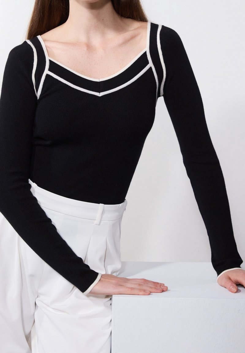 Femme portant un haut noir à manches longues avec bordure blanche et un pantalon blanc taille haute, reposant ses mains sur un bloc rectangulaire blanc.