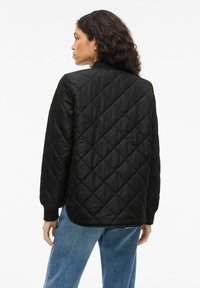 Črno prešito jakno z ovratnikom stoječega kroja, z diamantnim vzorcem in dolgimi rokavi, kombinirano z modrimi kavbojkami. Gladek tekstura.