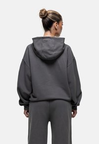Felpa oversize grigia scura con cappuccio, morbida al tatto, spalle cadenti e polsini a costine. Abbinata a pantaloni coordinati, design minimalista.