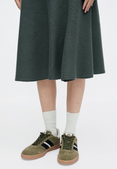 Ελίτ πράσινα σουέντ αθλητικά παπούτσια με μαύρες ρίγες, καουτσούκ σόλες, φορεμένα με λευκές κάλτσες μέχρι τον αστράγαλο και συνδυασμένα με μια σκούρα πράσινη φούστα.