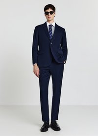 Abito blu navy con blazer aderente, rever a risvolto e tasche laterali, abbinato a una camicia a righe azzurre e una cravatta blu navy. Occhiali da sole neri e scarpe nere.