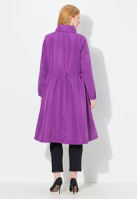 Manteau violet à manches longues avec une taille cintrée et un ourlet évasé, présentant un col haut et des poignets boutonnés, associé à des leggings noirs et des talons hauts.