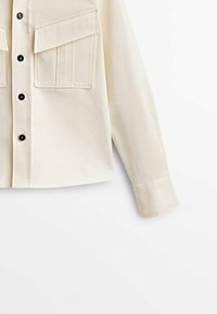 Chemise beige clair à manches longues, deux poches poitrine, devant à boutonner et boutons noirs. Texture lisse et design de col classique.