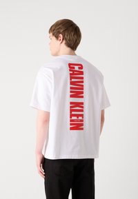 Hvit bomulls T-skjorte med en avslappet passform, med dristig rød "CALVIN KLEIN" skrift vertikalt på baksiden og en liten logo nedenfor.