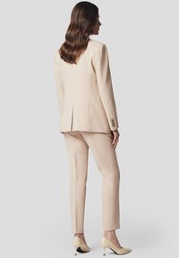 Beżowy garnitur skrojony na miarę z dopasowaną marynarką i wąskimi spodniami. Marynarka ma pojedynczy rozporek i detale w postaci guzików. Połączony z kremowymi szpilkami.