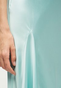 Main gauche reposant à côté d'une jupe en satin bleu clair avec un détail de couture et un pli doux.